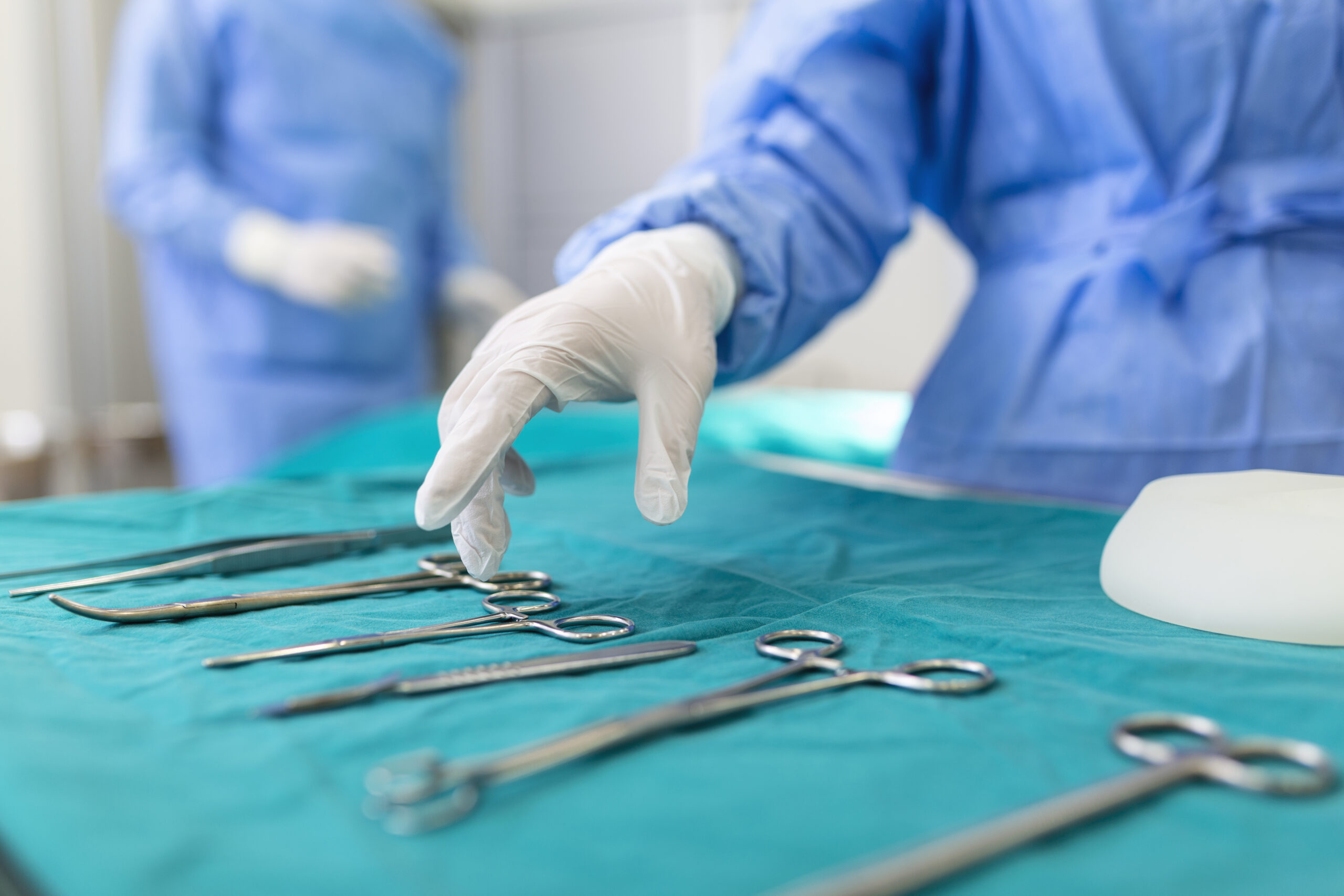 Nurse hand taking surgical instrument for group of surgeons at background operating patient in surgical theatre. Steel medical instruments ready to be used. Surgery and emergency concept